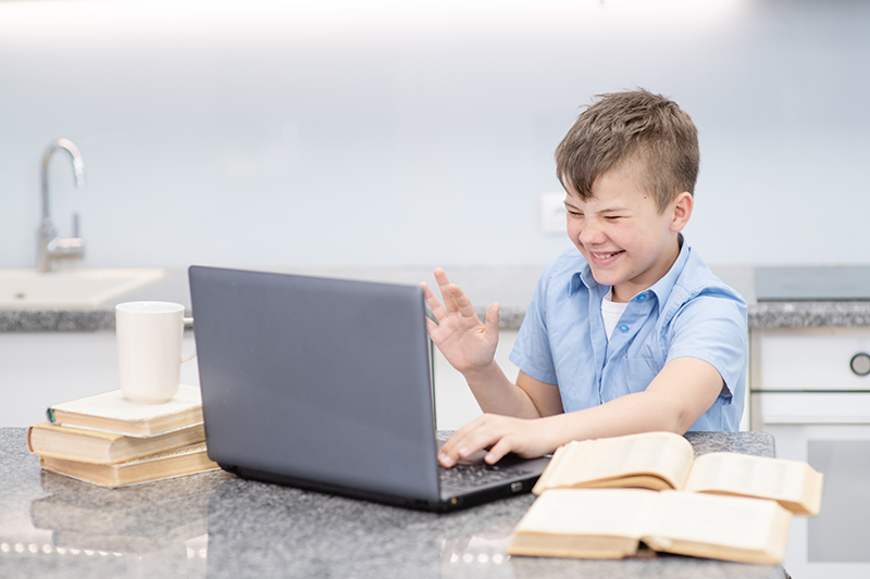 A boy is smiling while raising his hand while in a call through his remote learning lesson